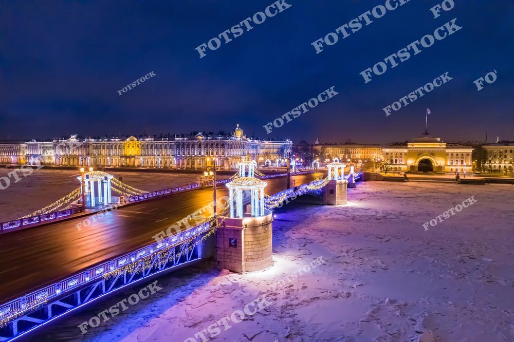 На фотографии изображён Дворцовый мост, украшенный праздничной иллюминацией, подчёркивающей его архитектурную изысканность. Вдали видны Зимний дворец, часть Эрмитажа, и Адмиралтейство, освещённые тёплым светом. Ледяная поверхность Невы и ночная атмосфера создают сказочное рождественское настроение. Этот снимок передаёт волшебство зимнего Санкт-Петербурга.
